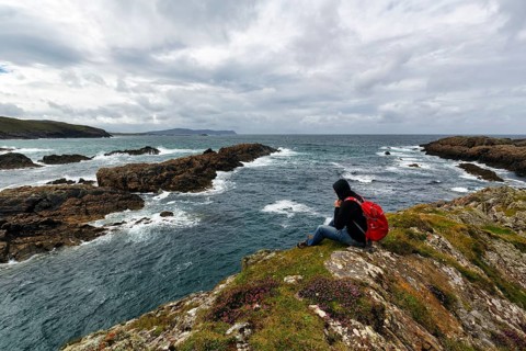 Hiker sitting on windy rocky coast, rain clouds, Melmore, Downings, Boyeeghter Strand, Murder Hole