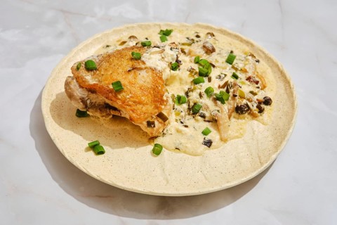 A plate holds a piece of cooked chicken alongside a creamy mushroom sauce. Chopped green onion is