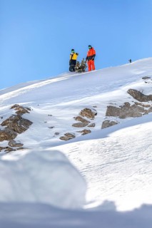 Two snowboarder friends in vibrant gear stand on a snowy mountain peak, poised to descend. Bright