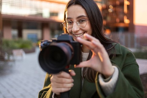 A smiling woman in a green coat holds a camera, capturing a moment outdoors at sunset She wears