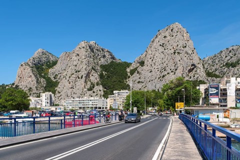 City road next to a bridge and high rocks with cars and buildings under bright blue summer sky,
