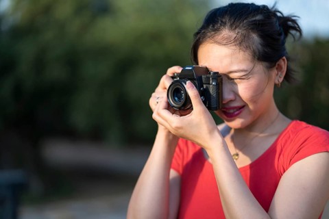 An Asian woman in a red shirt holds a vintage camera, capturing a picture outdoors. She focuses