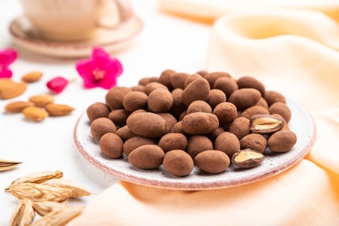 Almond in chocolate dragees on ceramic plate and a cup of coffee on white concrete background and