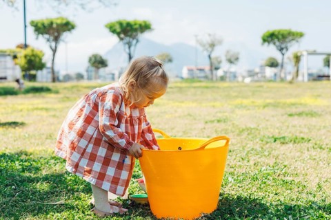 A young child in a checked dress bends over to look inside a large orange bucket on a grassy field