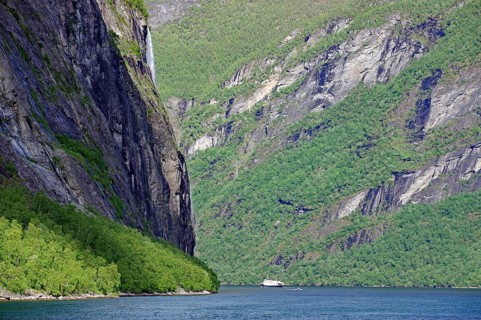A boat sails through a fjord surrounded by steep green cliffs under a blue sky with scattered