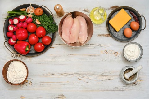 Fresh vegetables and raw chicken breasts are arranged on a wooden table. Red bell peppers,