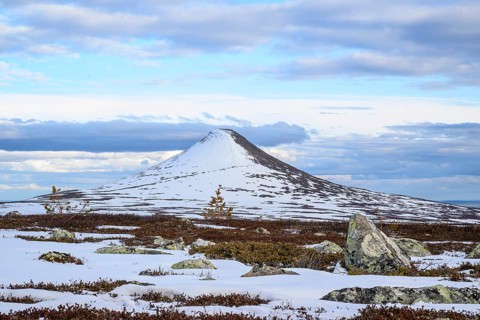 Snow-covered mountain under blue sky with clouds, surrounded by rocks and barren vegetation,