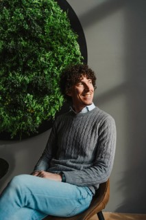 A young man with curly hair sits smiling beside a lush green vertical garden. The composition