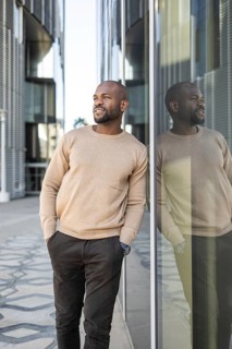 A cuban man in casual attire stands against a glass building, hands in pockets. He gazes
