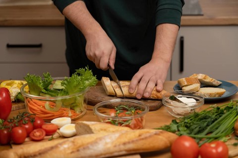 Hands cutting fresh bread amidst colorful vegetables like lettuce, carrots, and tomatoes. A