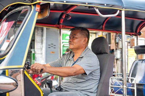 A Thai male tuk tuk driver sits in his vehicle, attentively awaiting passengers on a vibrant street