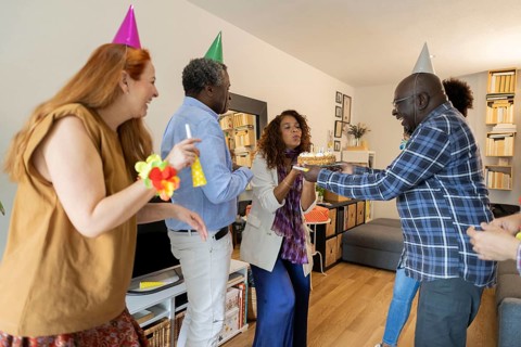 A lively indoor birthday celebration with friends. A woman joyfully blows out candles on a cake,