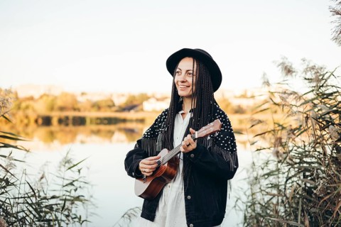 A woman with braided hair and a hat joyfully plays a ukulele near a serene lake during sunset,