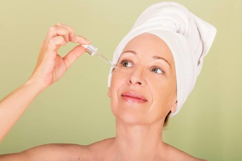 A woman with a towel wrapped around her head applies serum to her face, exemplifying a self-care