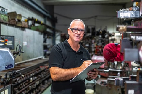 Mature male worker doing inventory in a metal cnc factory holding clipboard while looking at camera