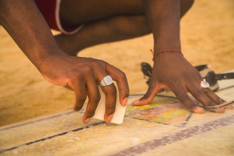 Person's hands waxing a surfboard on a sandy beach, preparing for a surfing session, focusing on