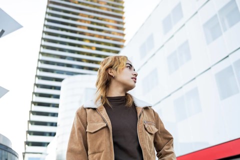 A Mixed-race woman in a brown jacket stands confidently in an urban setting, surrounded by modern