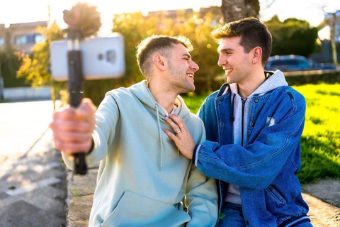 Smiling gay couple taking selfie outdoors using smartphone and gimbal, enjoying sunny day in urban