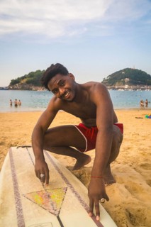 Young black man smiling at camera, crouching next to a surfboard on the sandy beach of la concha,