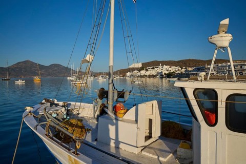 A sailboat in a quiet harbor with a view of white buildings and mountains in the background,