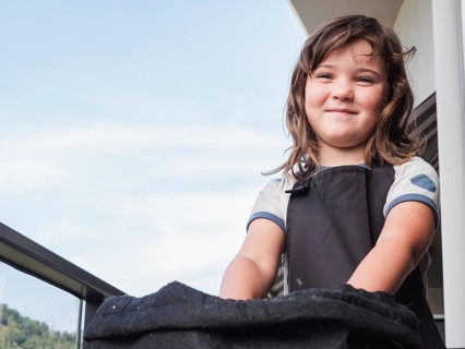 From below a young girl with a joyful expression stands in a balcony, dressed in casual attire and