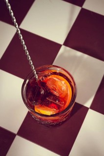 A drink fills a glass on a checkered table. Ice cubes and orange slices add color to the drink. A
