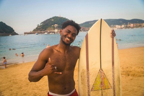 Young black man smiling and showing a shaka hand sign while standing with his surfboard on the