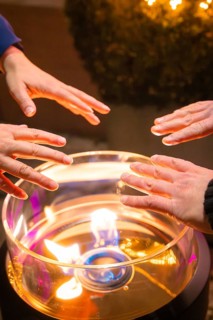 Hands warming themselves by a fire in a glass fixture, surrounded by warm lights at night,