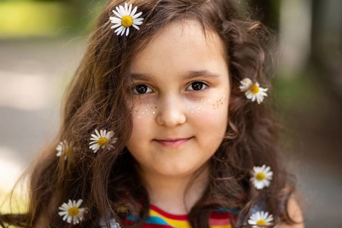 A happy little girl with daisies in her hair smiles warmly at the camera, embodying innocence and