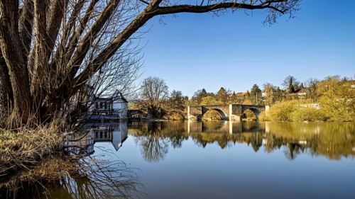 A peaceful river landscape with an old stone bridge and a clear sky, The Bartenwetz Stone Bridge in
