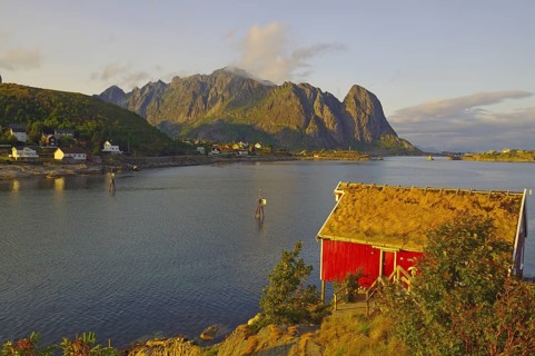 Red wooden hut with boat on the fjord shore, surrounded by mountains at sunset, Rorbuer, holiday