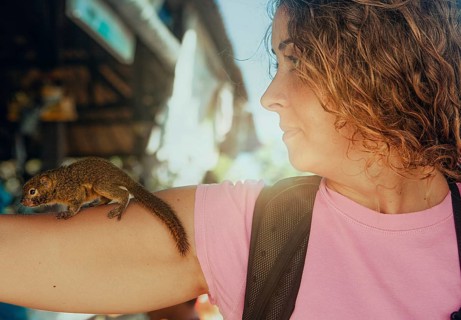 A woman watches a small tupaia perched on her arm in a bustling outdoor area in Bali. The image