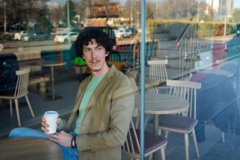 A businessman in casual attire sits by the window in a modern cafe, holding a takeaway coffee cup.