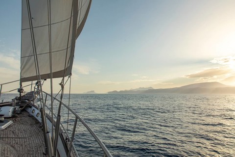 A sailboat gracefully cruises on the calm waters of the Mediterranean Sea, with the golden glow of