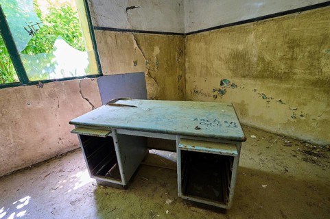 An abandoned room with an old desk, crumbling walls and damaged windows. Daylight falls through the