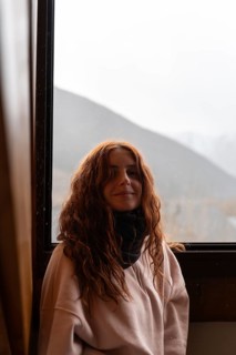 A young woman with red hair and a gentle smile is pictured comfortably inside during winter. She