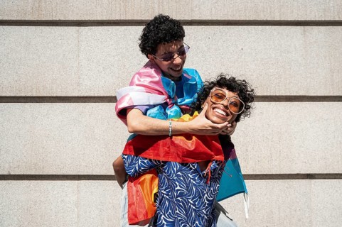 Two joyful people from a multiethnic LGBTQ+ couple embrace and laugh together, wrapped in colorful
