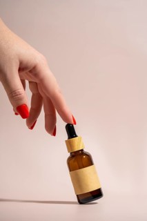A hand with vibrant red nails elegantly holds an cosmetic amber glass dropper bottle against a soft