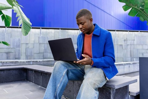Young African man with short hair sits on a bench, using a laptop in a modern outdoor setting with