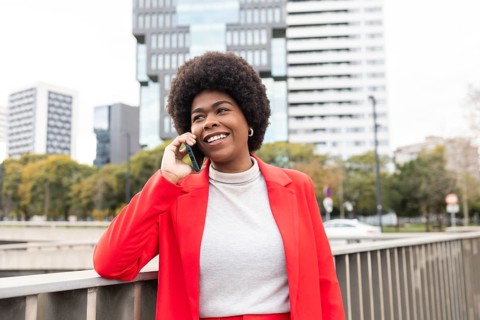 Smiling woman in a vibrant red jacket stands by an urban rail, chatting on her phone. Her confident