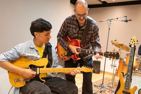 In a recording studio, a guitar instructor guides a student, who is holding a guitar, through a