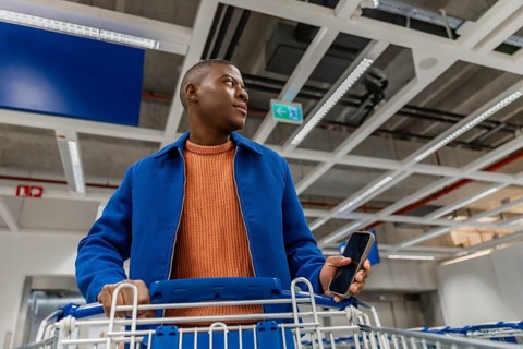 A young man stands in a busy store with a shopping cart. He holds a smartphone in one hand and