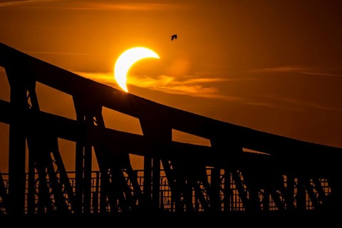 Partial solar eclipse above a large industrial bridge structure, creating a dramatic silhouette