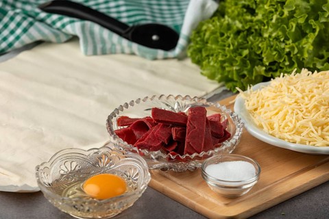 An egg and grated cheese together with puff pastry and ham on a wooden board
