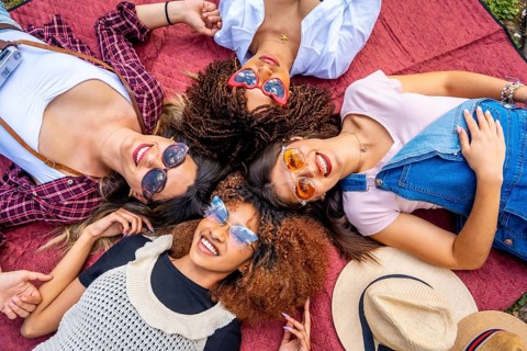 Four cheerful young women with sunglasses lying in circle on blanket enjoying sunny day in park