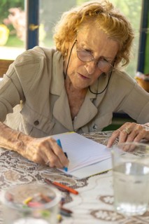 Senior woman with glasses drawing with crayons on a notebook at a table with a glass of water,