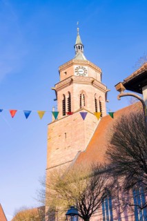 Historic church tower with clock in front of a bright blue sky, decorated with colourful flags,