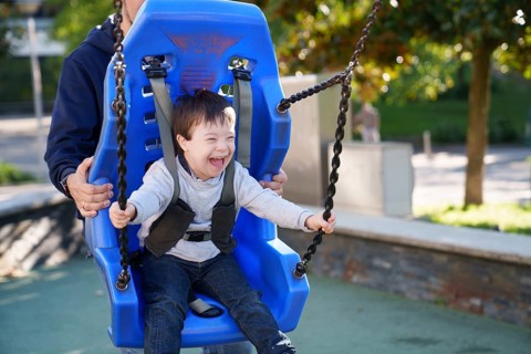 A child with Down syndrome happily swings on an adapted swing in a sunny park, supported by an
