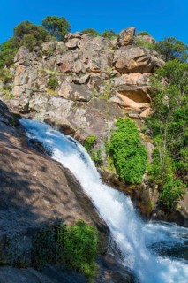 A mighty waterfall flows over large rocks in a forest landscape under a sunny sky, Cascada del