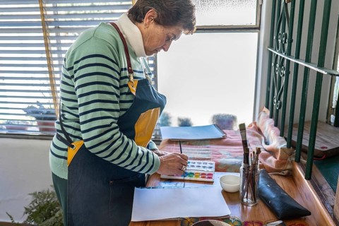 Older woman in denim apron stands over a wooden desk organizing watercolor supplies — palette,